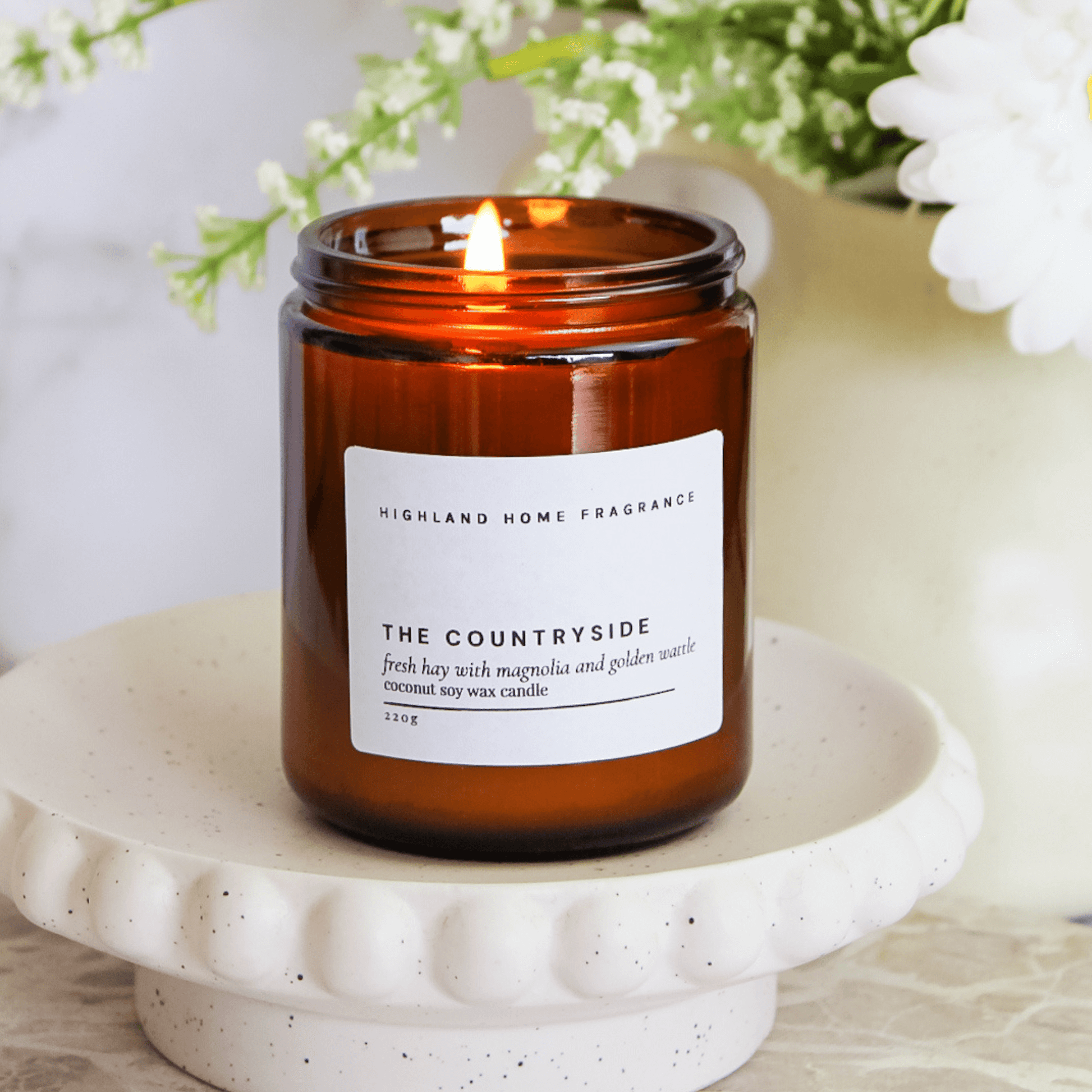 Highland Home Fragrance lit candle in the scent 'The Countryside' with notes for Florals and Fresh Hay. In an Amber jar with a lit wick.